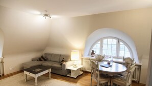 A loft-style apartment with a high ceiling, a cozy seating area with a sofa and a coffee table, a dining table with chairs, and a large arched window providing natural light.