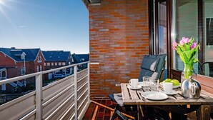 Un balcón con una mesa para dos, una silla, un florero y vistas a edificios al fondo.
