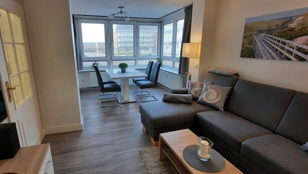 A modern living room with a sofa, a coffee table, and a dining area with chairs and a table. - By the sea - A modern and bright vacation apartment in the heart of Westerland. (Sylt)