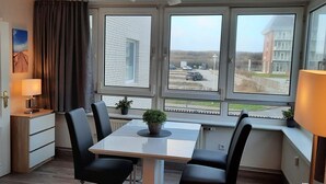 A compact room with a dining table, four chairs, a small kitchen area, and a window with a view of buildings and a road. - By the sea - A modern and bright vacation apartment in the heart of Westerland. (Sylt)