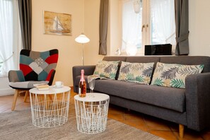 Ein Wohnzimmer mit einem grauen Sofa, einem gemusterten Sessel, zwei weißen Drahttischen, einer Champagnerflasche und einem Segelbootgemälde an der Wand.