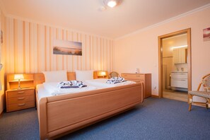A hotel room with a large bed, bedside tables, a chair, a wardrobe, and a bathroom visible through an open door. - Kim in Haus Wega - Relaxed island life: Cozy vacation apartment in (Sylt)