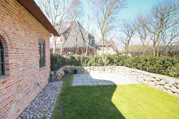 Une maison en briques avec un chemin de pierres menant à une cour verdoyante, entourée d’arbres et d’un mur de pierres.