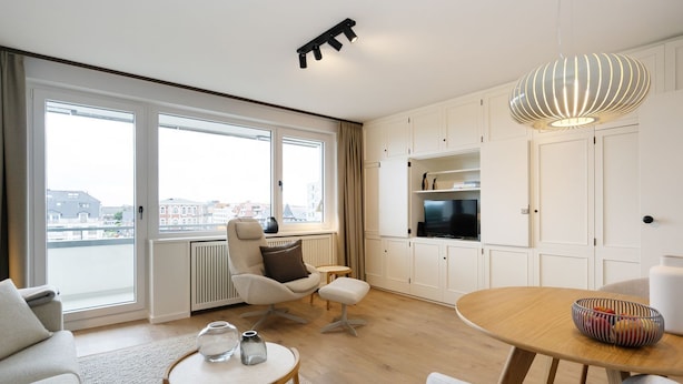 Un salon moderne avec un canapé, une table ronde, un fauteuil, un téléviseur et une grande fenêtre donnant sur la ville.