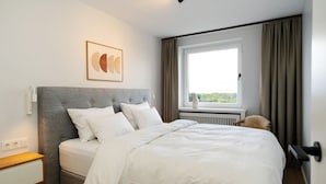 Un lit bien fait, avec une literie blanche, une tête de lit grise, une table de chevet avec une lampe et une fenêtre avec des rideaux.