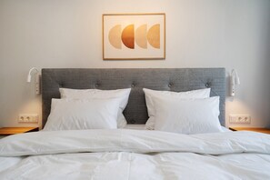 Ein ordentlich bezogenes Bett mit weißer Bettwäsche, einem grauen gepolsterten Kopfteil und einem gerahmten Kunstwerk darüber.