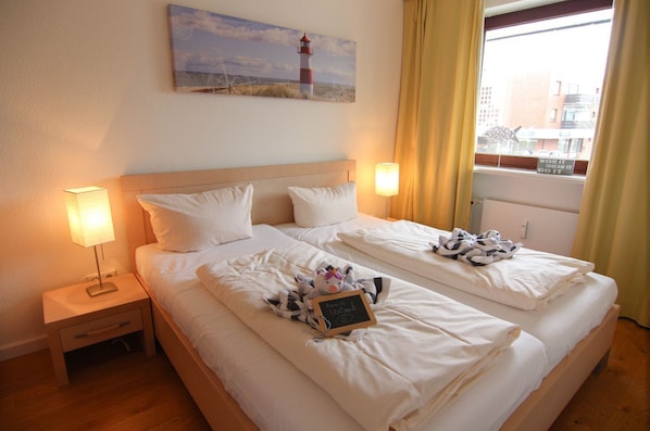A hotel room with two beds, a nightstand with a lamp, a painting of a lighthouse, and a window with curtains.