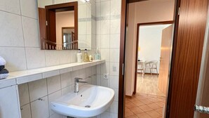 Ein Badezimmer mit einem weißen Waschbecken, einem Spiegel und einem gefliesten Boden. Eine Tür führt in einen Raum mit einem Sessel und einem Tisch.