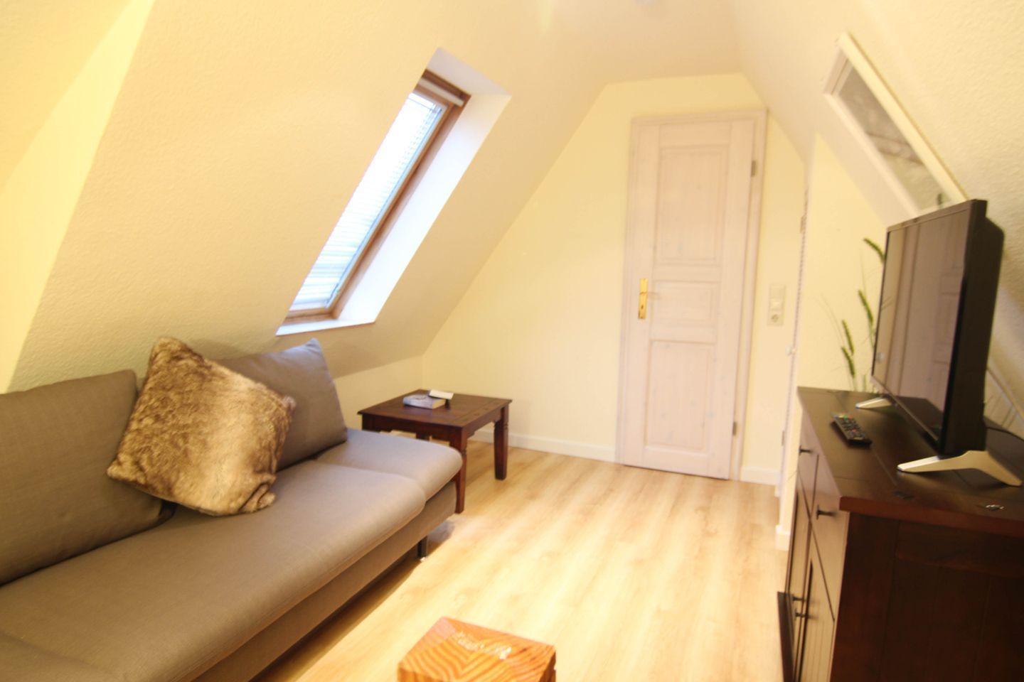 A small, cozy room with a sloped ceiling, a sofa, a small table, a TV, and a door.