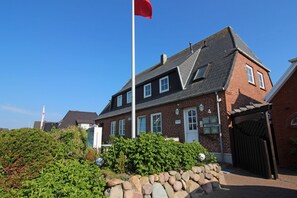 A two-story brick house with a red flag on a flagpole, a stone wall, and a garden with green shrubs and a stone pathway.