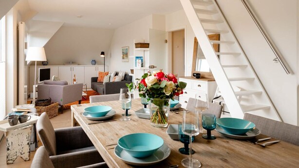 Un salon moderne avec un coin repas, comprenant une table à manger en bois, dressée avec de la vaisselle bleue et des verres, un canapé et un escalier.