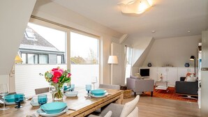 Un espace de repas moderne avec une table en bois dressée pour quatre personnes, de grandes fenêtres et une cuisine en arrière-plan.