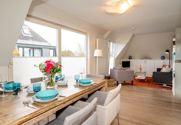 A modern dining area with a wooden table set for four, large windows, and a kitchen in the background.