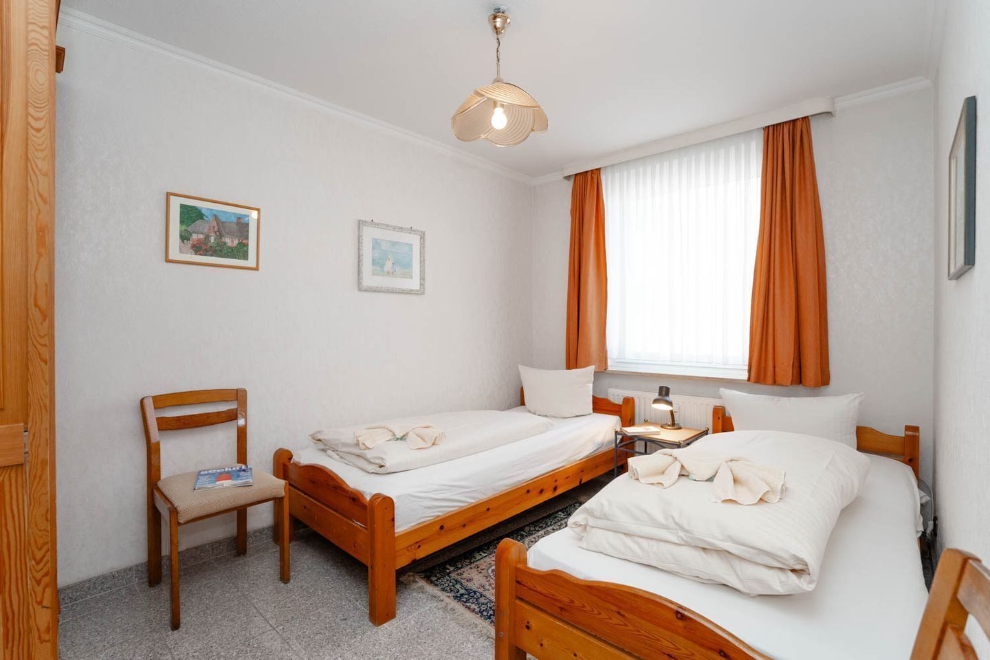 A room with two wooden beds, a chair, a table with a lamp, and two framed pictures on the wall.
