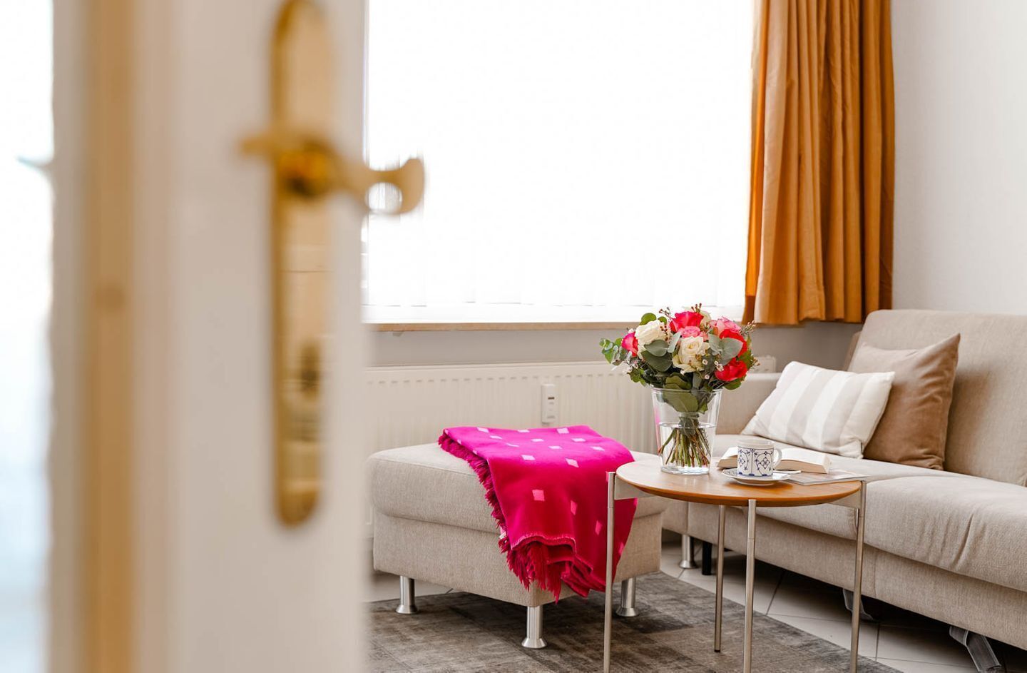 Ein modernes Wohnzimmer mit einer Couch, einem kleinen Tisch mit Blumenvase und einer rosafarbenen Decke auf einem Pouf.
