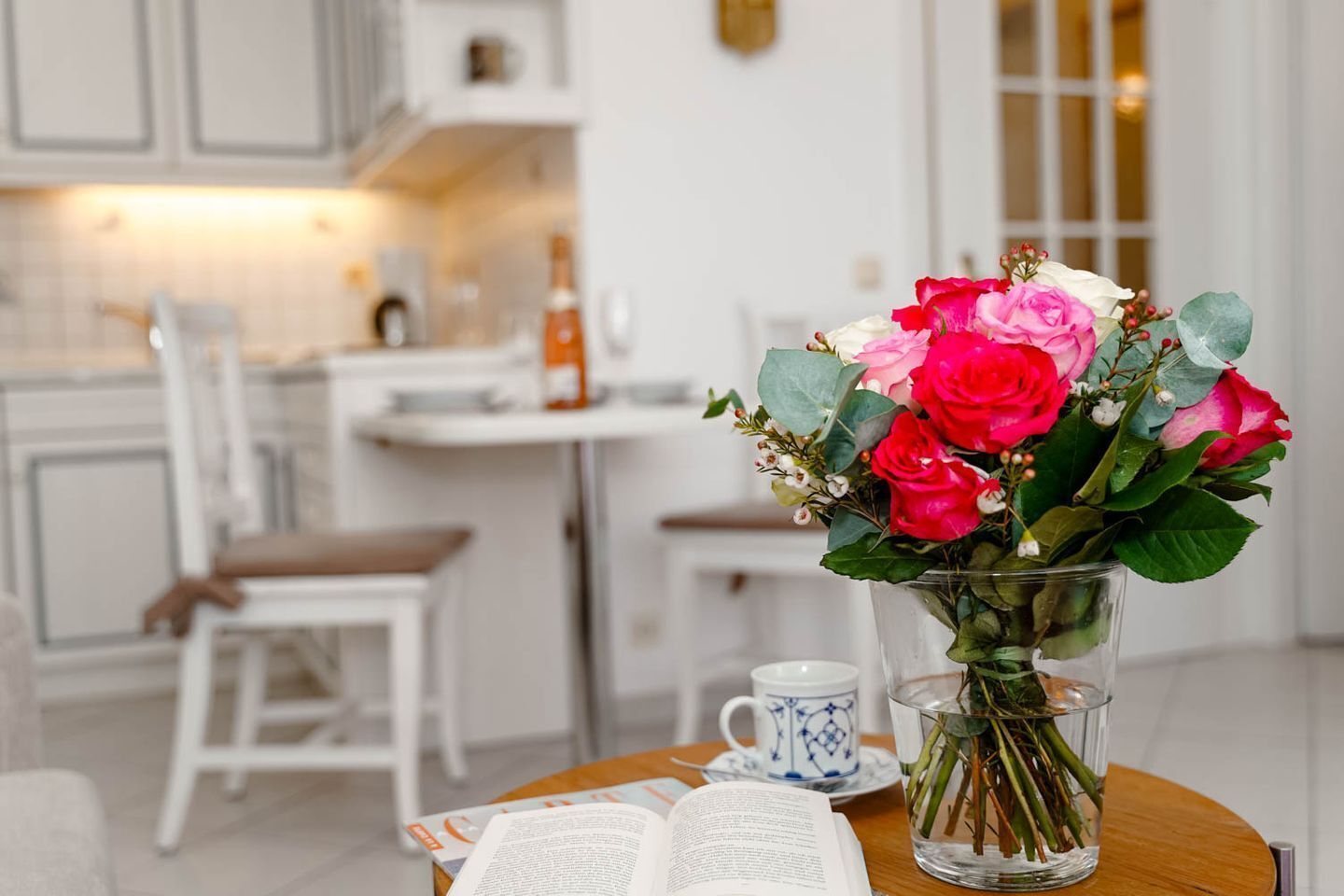 Une cuisine moderne avec des meubles blancs, un coin repas avec une table dressée pour deux personnes et un vase de fleurs sur la table.