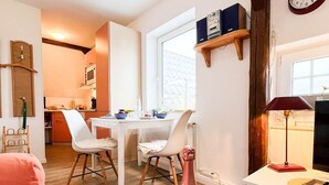 A cozy room with a bed, a dining area with chairs, a small table with a bottle and an apple, and a clock on the wall. - East - Relaxed island life: Cozy vacation apartment in Westerland on Sylt (Sylt)