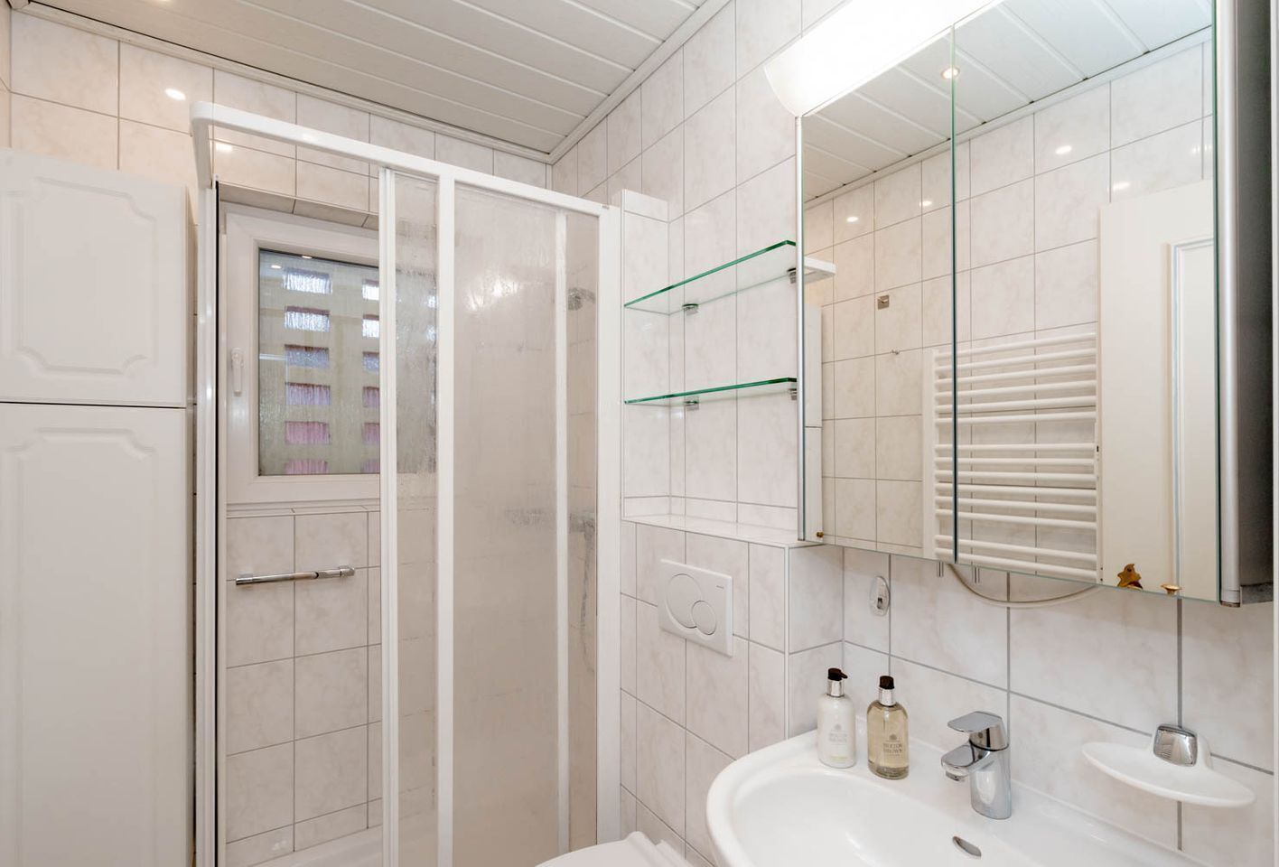 A modern bathroom with a glass shower enclosure, a white sink with two bottles of liquid soap, and a mirror above the sink.