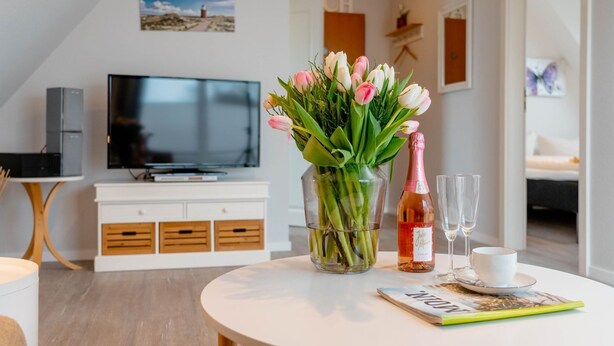 Un salon moderne avec un téléviseur à écran plat, un meuble blanc, une table ronde avec un magazine, un vase de tulipes, une bouteille de champagne et deux verres.