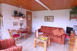 Un salon chaleureux avec un canapé, des fauteuils, une table basse sur laquelle sont disposés des fruits, un téléviseur et un plafond en bois.