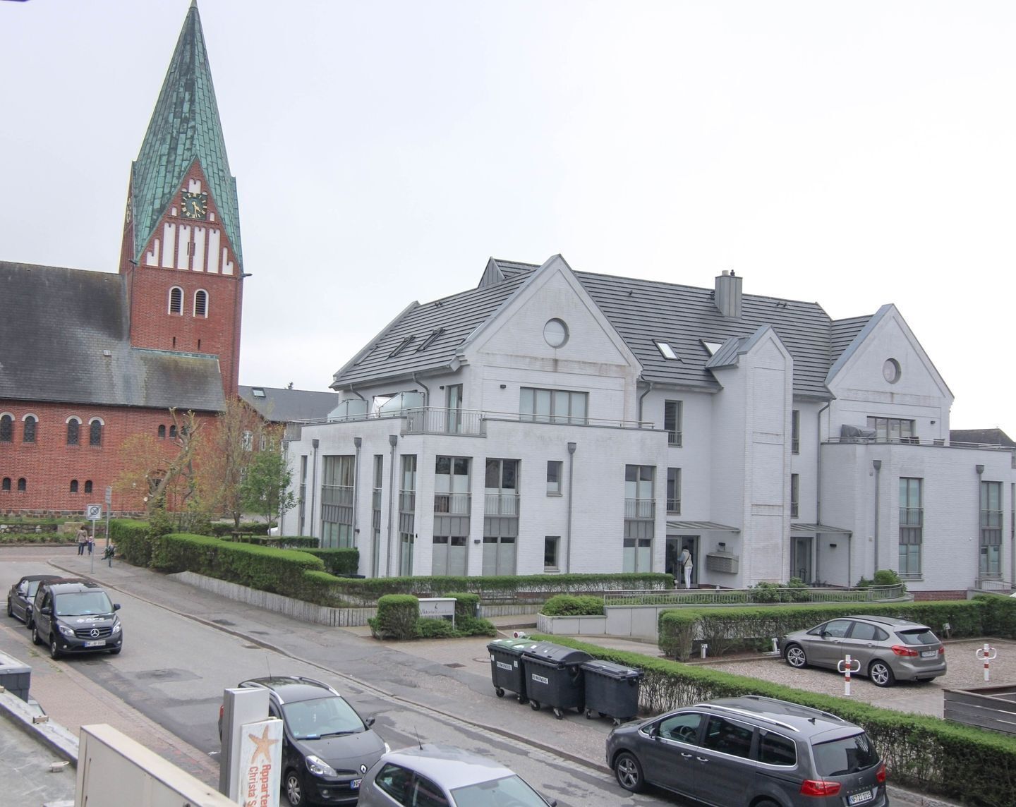 A modern building with a large clock tower, a parking area with several cars, and a well-maintained garden.