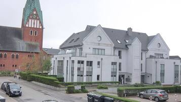 A modern building with a large clock tower, a parking area with several cars, and a well-maintained garden.