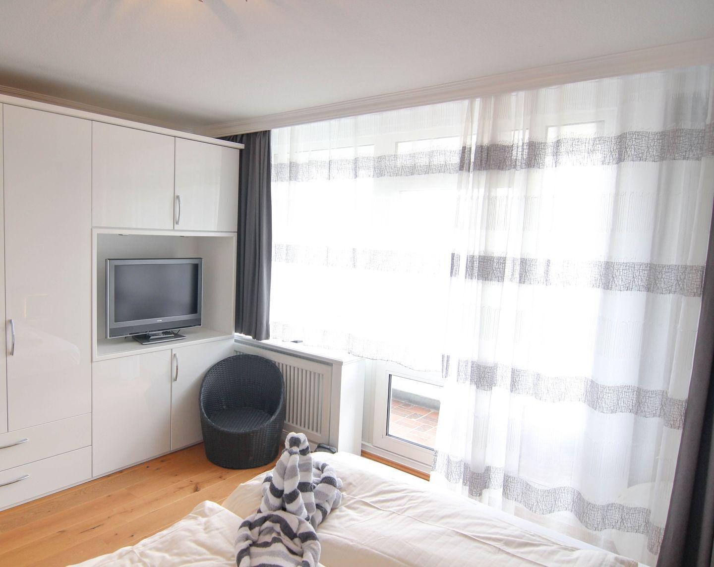 Une chambre d’hôtel moderne dotée d’un téléviseur à écran plat fixé à un meuble blanc, d’une grande fenêtre avec des rideaux légers, d’un fauteuil en osier et d’un lit recouvert d’une couverture à rayures.