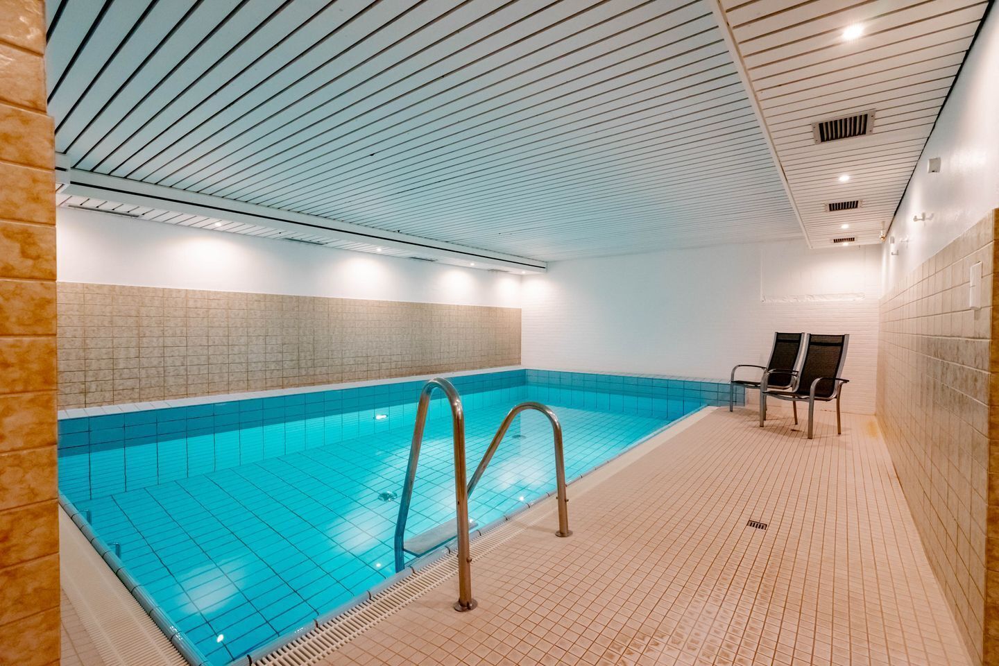 An indoor swimming pool with a tiled floor and walls, a metal pool ladder, and two chairs near the wall.