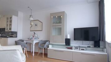 En moderne stue med sofa, sofabord i glas, fjernsyn og et køkkenområde med skabe samt et spisebord.
