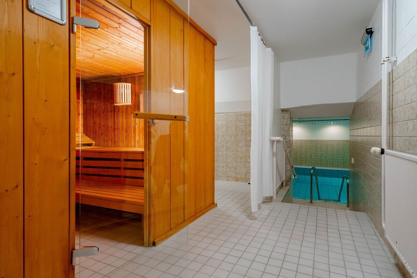 A sauna room with wooden walls and a glass door, leading to a tiled bathroom with a blue-tiled shower area.