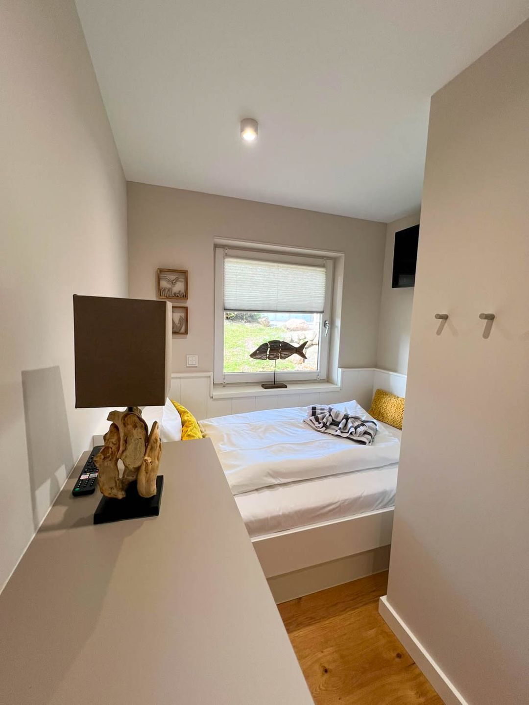 Un dormitorio con cama, ventana con persianas, mesita de noche con lámpara y televisor montado en la pared.
