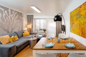 Ein modernes Wohnzimmer mit einem grauen Sofa, gelben Kissen, einem Holzesstisch mit Tellern und Gläsern, einer schwarzen Stehlampe und einem farbenfrohen abstrakten Gemälde an der Wand.