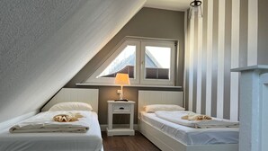 Une petite chambre mansardée, bien éclairée, avec deux lits simples, une table de chevet et une fenêtre laissant entrer la lumière naturelle.