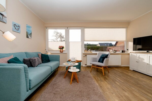 A modern living room with a teal sofa, a coffee table, a chair, and a TV. There are windows with blinds, a radiator, and a view of buildings outside. - Alea - A modern and bright vacation apartment in the heart of Westerland. (Sylt)