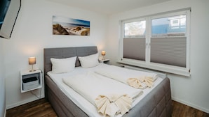 A bedroom with a bed, bedside tables, a window with blinds, a painting on the wall, and a ceiling light fixture. - Alea - A modern and bright vacation apartment in the heart of Westerland. (Sylt)