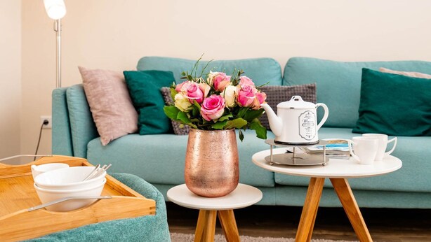 A cozy living room with a teal sofa, a copper vase with pink flowers, a white teapot, and a tray with bowls and utensils. - Alea - A modern and bright vacation apartment in the heart of Westerland. (Sylt)