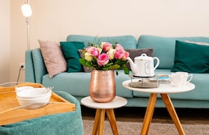 A cozy living room with a teal sofa, a copper vase with pink flowers, a white teapot, and a tray with bowls and utensils.