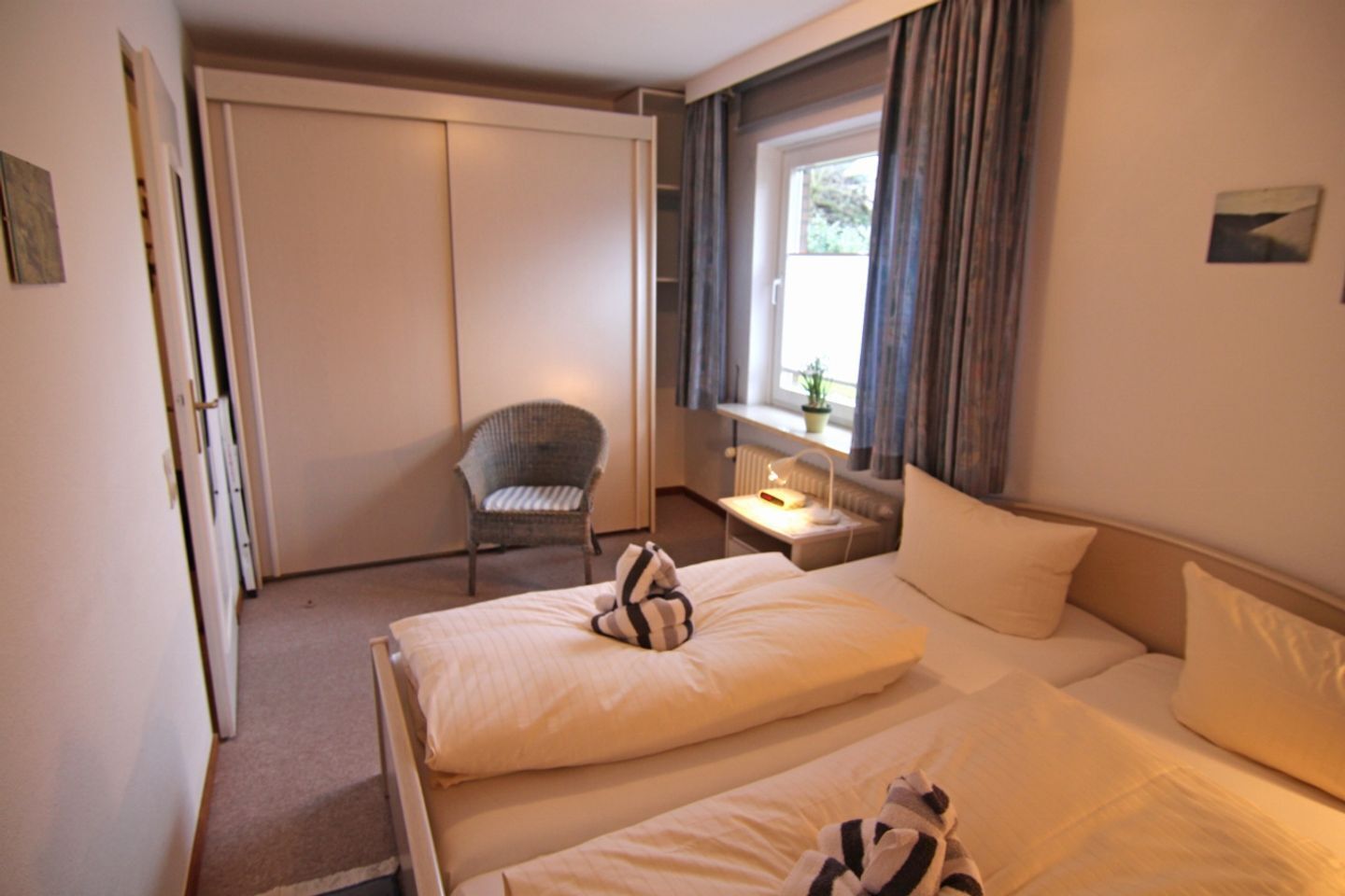 A hotel room with two beds, a chair, a window with curtains, and a small table with a lamp.