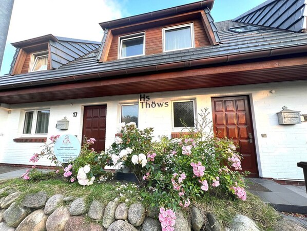 A two-story building with a sign reading "Hs Thöws" and a stone wall with flowers in front.