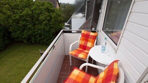 Un balcon avec deux chaises blanches et des coussins orange, une petite table ronde et une bouteille d’eau.