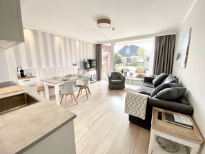 A modern apartment with a kitchen, dining area, and living room. It features a large window, a sofa, and a TV.