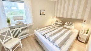 A bedroom with a large bed, a chair, a radiator, a window with curtains, a bedside table with a lamp, and a wall clock.