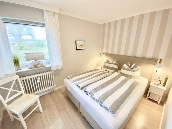 A bedroom with a large bed, a chair, a radiator, a window with curtains, a bedside table with a lamp, and a wall clock.