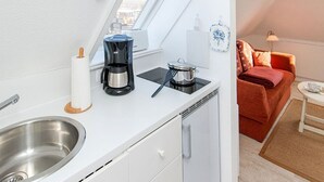 Une petite cuisine équipée avec une cuisinière, une machine à café et un évier. Une petite fenêtre laisse entrer la lumière naturelle, et un canapé rouge est visible dans le salon adjacent.