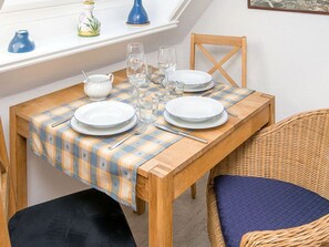 Une table à manger est dressée avec des assiettes, des verres et des couverts. On y trouve une chaise en osier avec un coussin, une fenêtre donnant sur un beau paysage et une petite table ornée d’objets décoratifs.