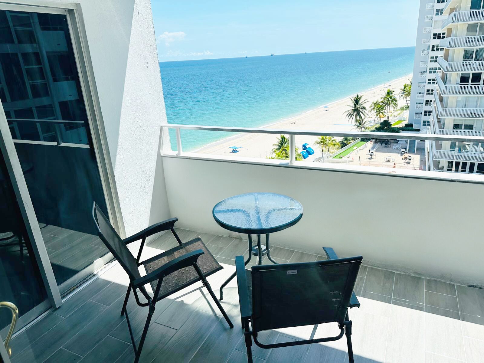 A balcony with a glass table and two chairs, overlooking a beach and ocean.