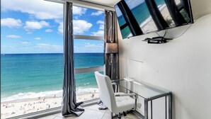 A room with a large window overlooking a beach, a flat-screen TV mounted on the wall, and a white chair. - Beachfront condo-We are on the Beach-Views every window-Patio Pool-No resort fee (Fort Lauderdale)