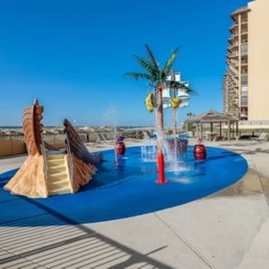A water play area with a slide and water jets, surrounded by a pool and a building.
