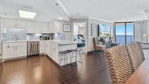 A modern kitchen with white cabinets, a dishwasher, and a large island with stools. There is a balcony with a view of the ocean.