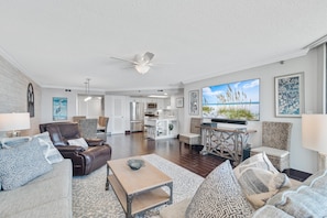 A spacious living room with a large flat-screen TV, comfortable seating, a dining area, and a kitchen in the background.